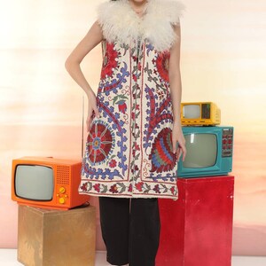 May include: A long white vest with a off white fur collar and colorful floral embroidery. The vest is worn over black pants and black heels.