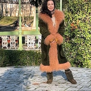 May include: A woman wearing a green velvet coat with a long, fluffy pink fur collar and cuffs. She is standing in front of a green gazebo with a white wrought iron railing. The ground is made of stone pavers.