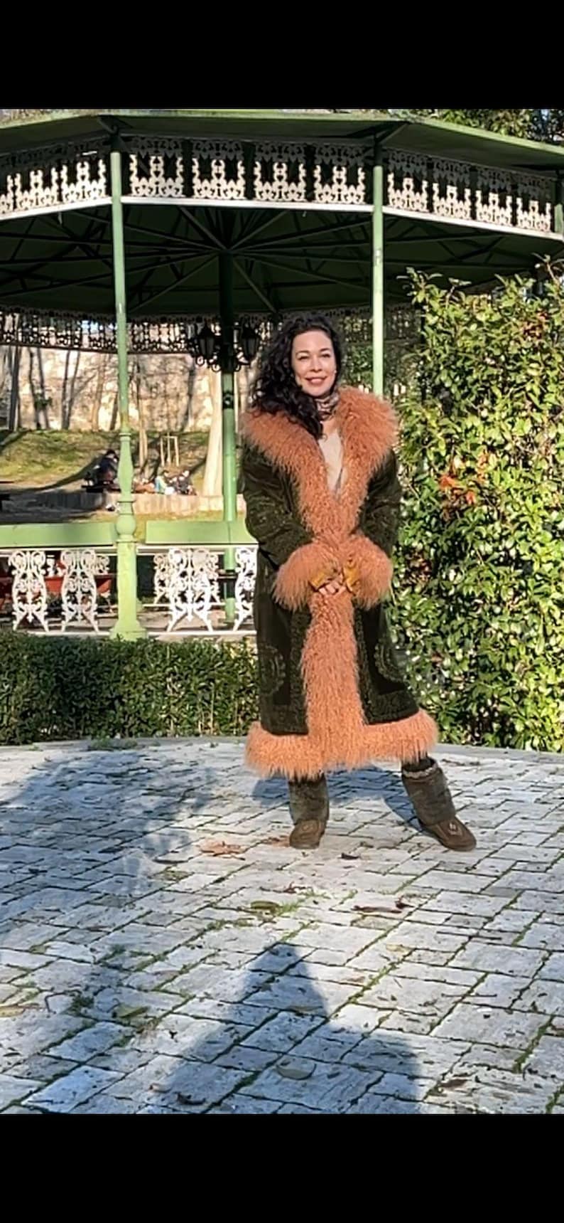 May include: A woman wearing a green velvet coat with a long, fluffy pink fur collar and cuffs. She is standing in front of a green gazebo with a white wrought iron railing. The ground is made of stone pavers.