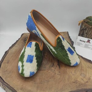 May include: A pair of green, blue, and white patterned velvet slippers with brown leather trim. The slippers are on a wooden surface.