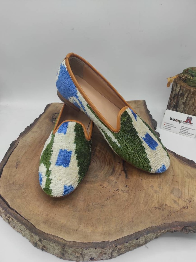 May include: A pair of green, blue, and white patterned velvet slippers with brown leather trim. The slippers are on a wooden surface.