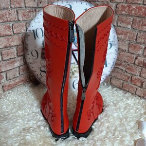 Puede incluir: Un par de botas de cuero rojo vibrante con intrincados bordados florales. Las botas tienen cierre de cremallera en la parte trasera. Las botas est&aacute;n sobre un fondo texturizado. Dise&ntilde;o cl&aacute;sico con un toque moderno.