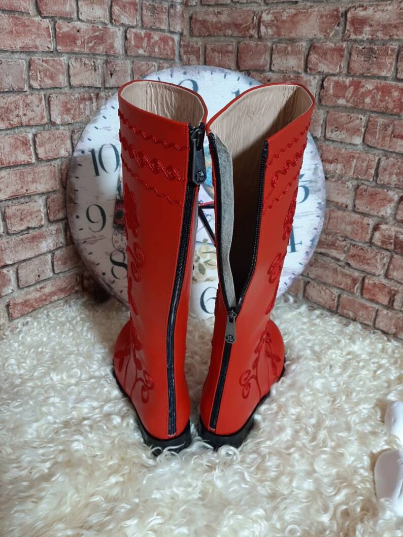 Puede incluir: Un par de botas de cuero rojo vibrante con intrincados bordados florales. Las botas tienen cierre de cremallera en la parte trasera. Las botas est&aacute;n sobre un fondo texturizado. Dise&ntilde;o cl&aacute;sico con un toque moderno.