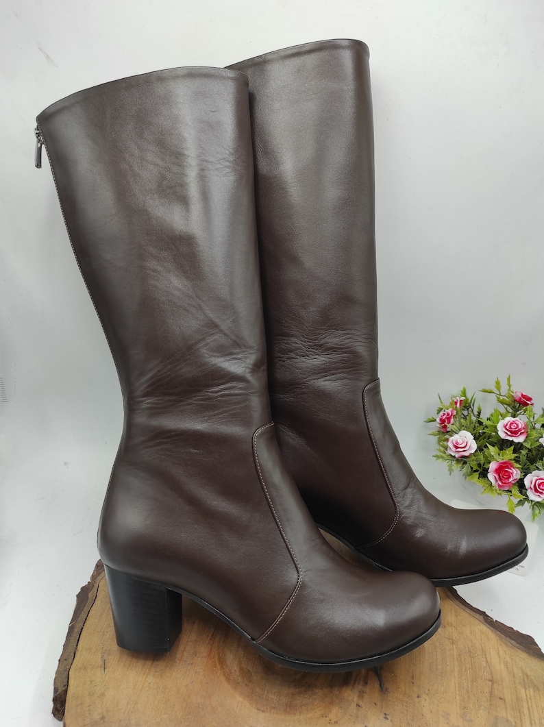 May include: A pair of brown leather knee-high boots with a block heel. The boots have a zipper on the back and a pointed toe.