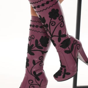 Puede incluir: Un par de botas hasta la rodilla de color ciruela intenso con patrones florales bordados en negro y dise&ntilde;os de enredaderas. Las botas tienen una punta redondeada, un tac&oacute;n de bloque resistente y l&iacute;neas onduladas decorativas en la parte superior.