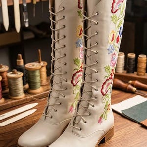 May include: A pair of cream-colored, knee-high boots with intricate floral embroidery in red, pink, yellow, and blue. The boots have a lace-up design and a low heel, showcasing a blend of style and craftsmanship.