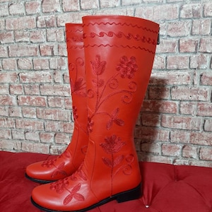 Puede incluir: Un par de botas rojas altas hasta la rodilla. Las botas presentan intrincados bordados florales en hilo rojo a juego. El dise&ntilde;o incluye un patr&oacute;n cosido decorativo en la parte superior y un cierre de cremallera. Las botas se exhiben sobre una superficie acolchada roja.
