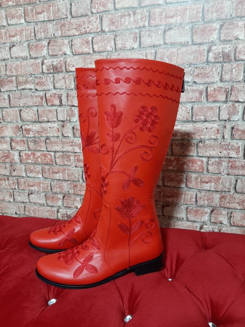 Puede incluir: Un par de botas rojas altas hasta la rodilla. Las botas presentan intrincados bordados florales en hilo rojo a juego. El dise&ntilde;o incluye un patr&oacute;n cosido decorativo en la parte superior y un cierre de cremallera. Las botas se exhiben sobre una superficie acolchada roja.