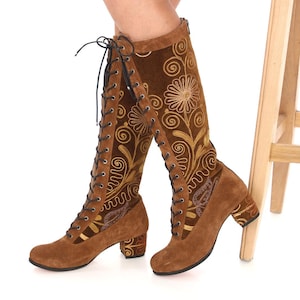 May include: Brown suede knee-high boots with a block heel and gold floral embroidery. The boots have a lace-up front and a zipper on the back.