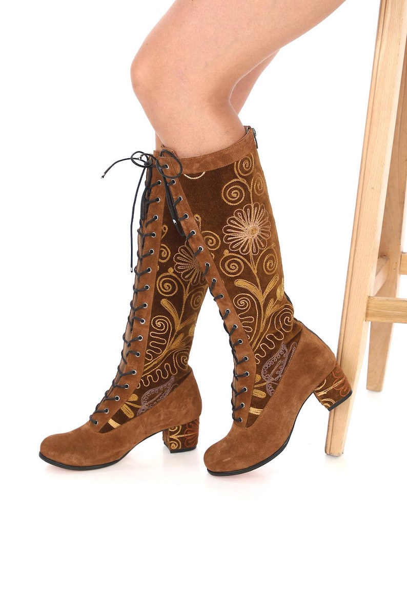 May include: Brown suede knee-high boots with a block heel and gold floral embroidery. The boots have a lace-up front and a zipper on the back.