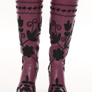 Puede incluir: Botas hasta la rodilla en color morado ciruela intenso con intrincados bordados negros. El dise&ntilde;o incluye motivos florales y patrones de remolinos que se extienden desde la punta hasta la parte superior de la ca&ntilde;a, con l&iacute;neas onduladas.