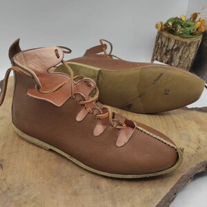 May include: A pair of brown leather sandals with laces. The sandals have cutouts on the top and a closed toe. The sandals are on a wooden surface.