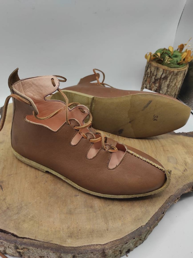 May include: A pair of brown leather sandals with laces. The sandals have cutouts on the top and a closed toe. The sandals are on a wooden surface.