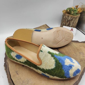 May include: A pair of green, blue, and white patterned velvet slippers with brown leather trim. The slippers have a brown leather sole.