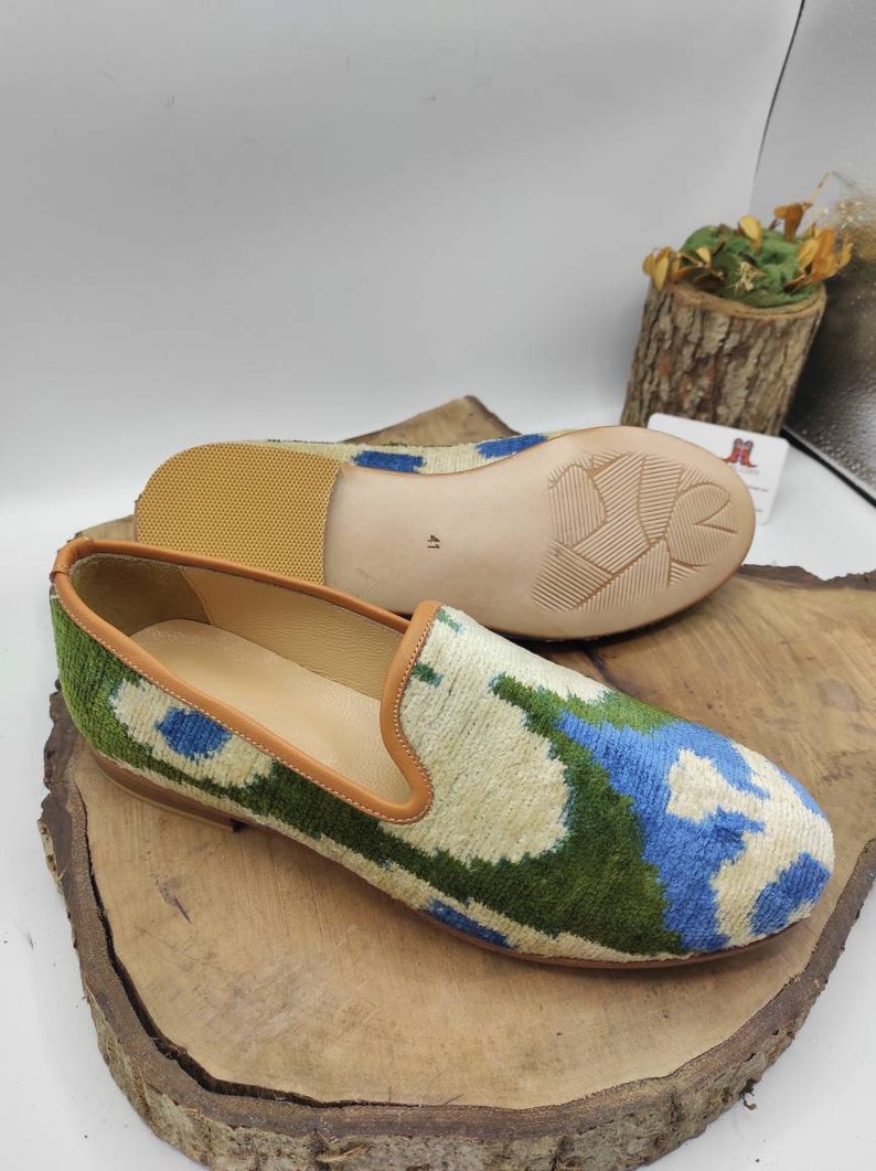 May include: A pair of green, blue, and white patterned velvet slippers with brown leather trim. The slippers have a brown leather sole.