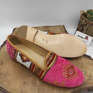 May include: A pair of pink and patterned slip-on shoes with a leather sole. The shoes have a geometric design in shades of red, yellow, and white. The size is 39. The shoes are displayed on a wooden surface.