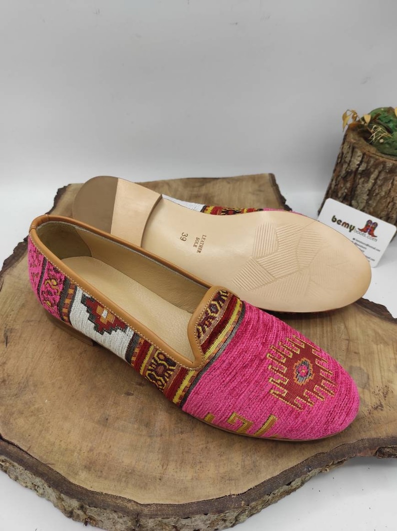 May include: A pair of pink and patterned slip-on shoes with a leather sole. The shoes have a geometric design in shades of red, yellow, and white. The size is 39. The shoes are displayed on a wooden surface.