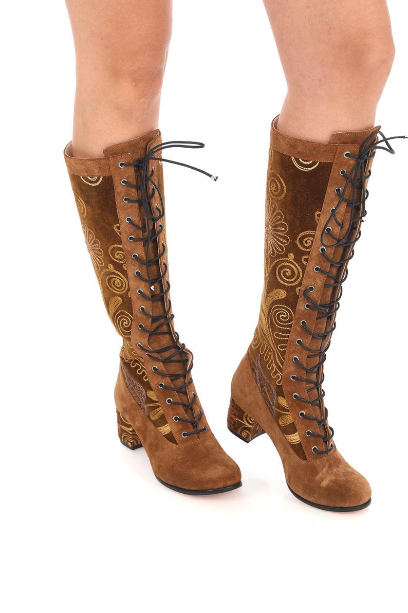May include: Brown suede knee-high boots with gold embroidery and black laces. The boots have a block heel.