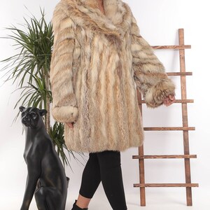 May include: A long, light brown fur coat with a wide collar and cuffs. The coat is worn over black leggings and black ankle boots. A black panther statue and a wooden ladder are in the background.