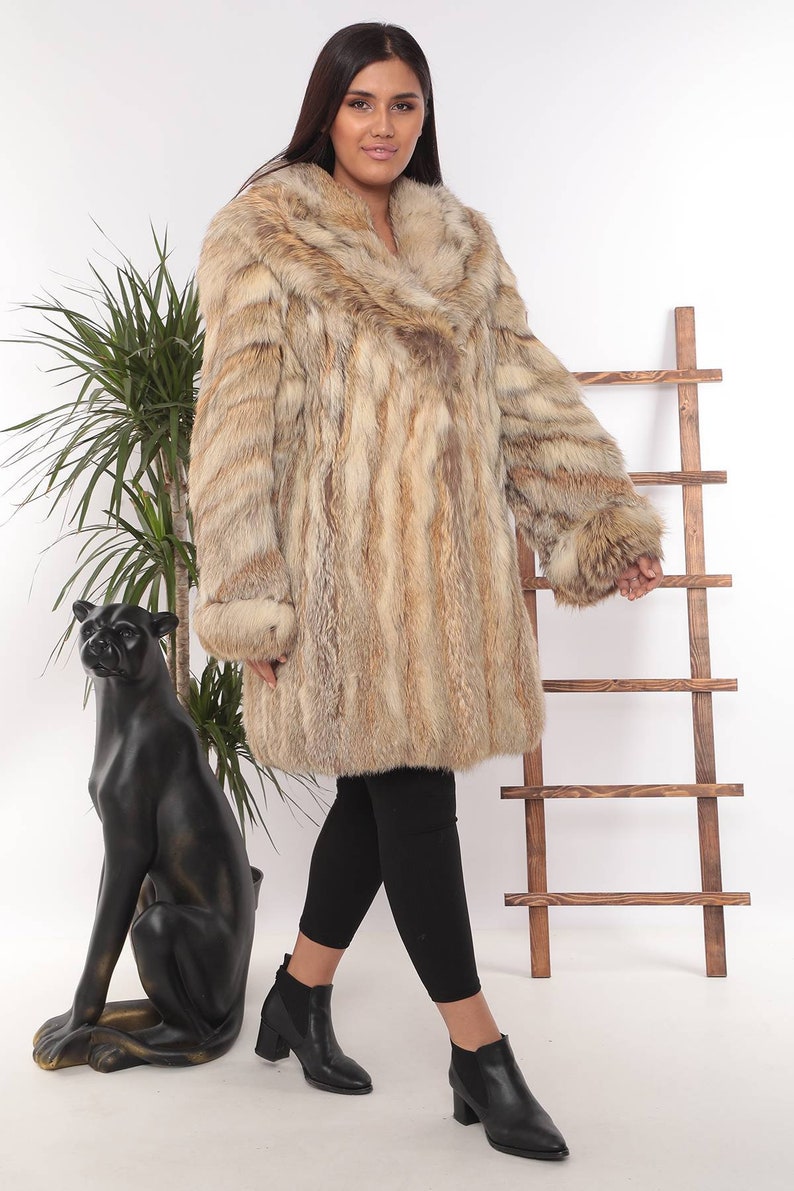 May include: A long, light brown fur coat with a wide collar and cuffs. The coat is worn over black leggings and black ankle boots. A black panther statue and a wooden ladder are in the background.
