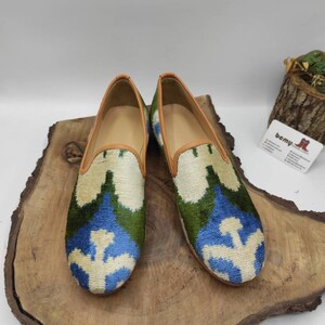 May include: A pair of blue, green, and white patterned velvet loafers with a brown leather trim. The loafers are sitting on a wooden surface.