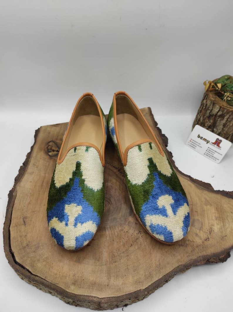 May include: A pair of blue, green, and white patterned velvet loafers with a brown leather trim. The loafers are sitting on a wooden surface.