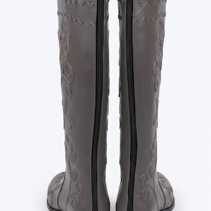 Puede incluir: Un par de botas grises hasta la rodilla con bordados florales. Las botas tienen una cremallera en la parte trasera y un tac&oacute;n peque&ntilde;o y plano. Las costuras decorativas y el bordado a&ntilde;aden un toque de elegancia.