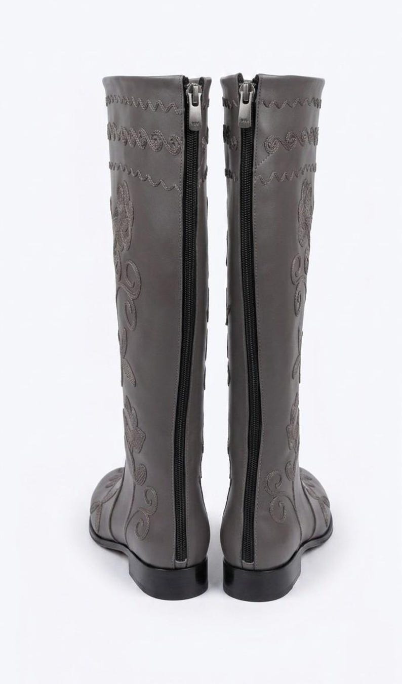 Puede incluir: Un par de botas grises hasta la rodilla con bordados florales. Las botas tienen una cremallera en la parte trasera y un tac&oacute;n peque&ntilde;o y plano. Las costuras decorativas y el bordado a&ntilde;aden un toque de elegancia.