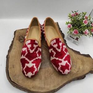 May include: A pair of red and white patterned velvet loafers with a brown leather trim. The loafers are on a wooden surface.