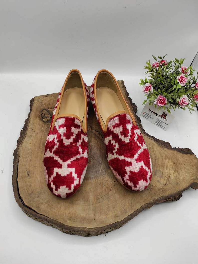 May include: A pair of red and white patterned velvet loafers with a brown leather trim. The loafers are on a wooden surface.