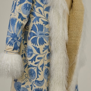 May include: A long white and blue embroidered coat with a cream fur trim. The coat is lined with a tan fabric.
