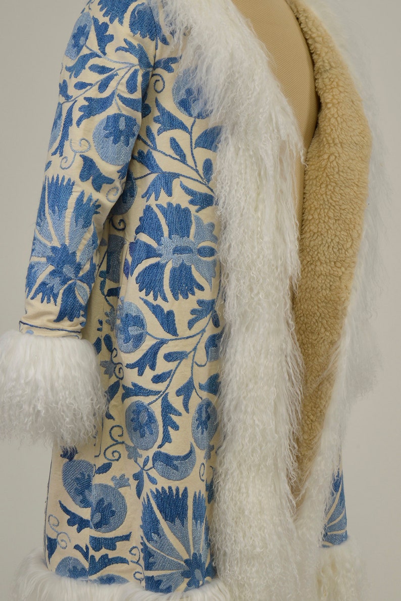 May include: A long white and blue embroidered coat with a cream fur trim. The coat is lined with a tan fabric.