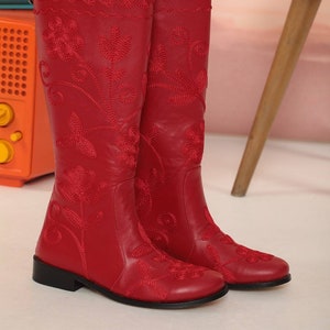 Puede incluir: Un par de botas rojas de cuero hasta la rodilla con intrincados bordados florales. Las botas tienen un tac&oacute;n bajo y un ribete bordado decorativo en la parte superior. Las botas se exhiben sobre una superficie blanca.