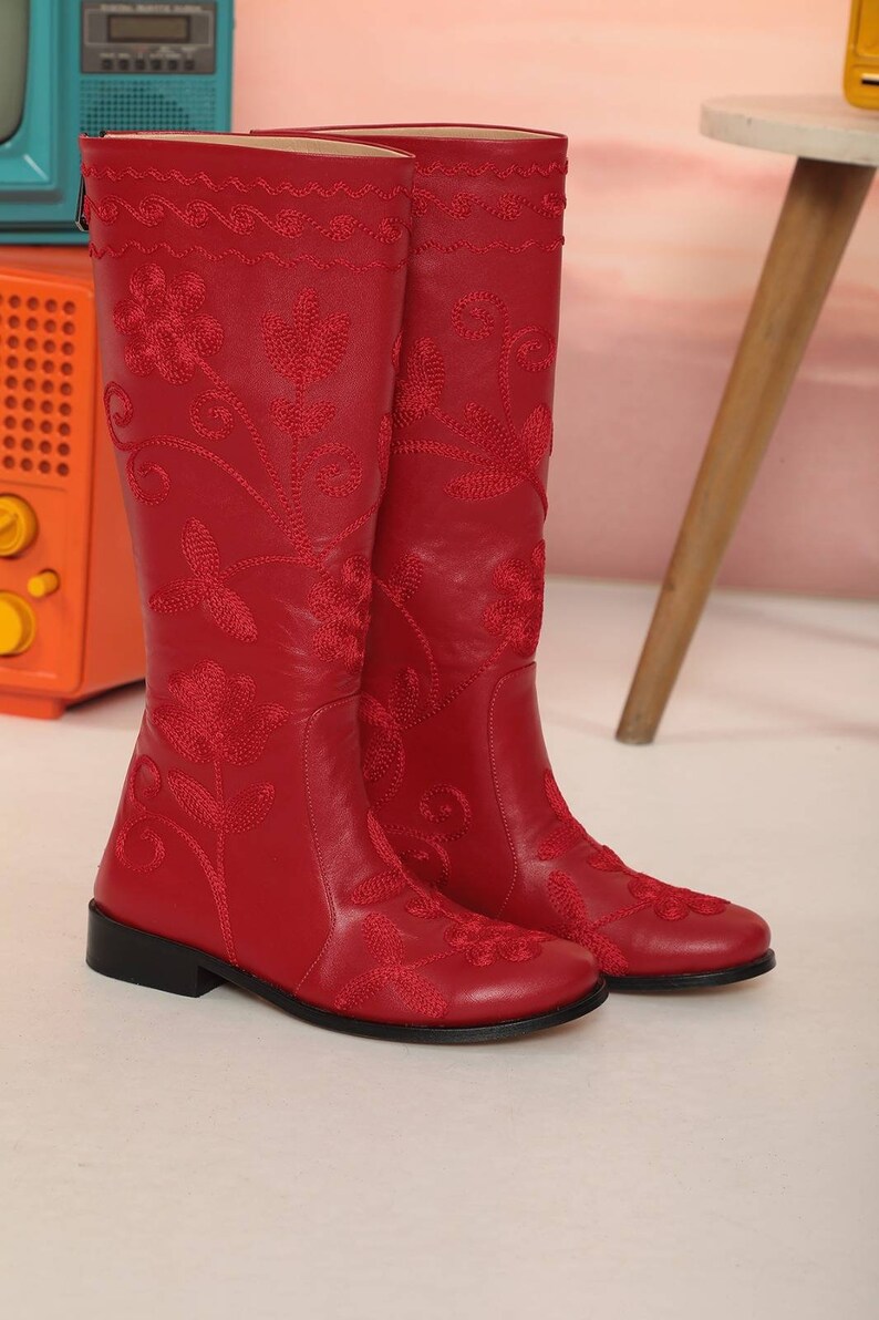 Puede incluir: Un par de botas rojas de cuero hasta la rodilla con intrincados bordados florales. Las botas tienen un tac&oacute;n bajo y un ribete bordado decorativo en la parte superior. Las botas se exhiben sobre una superficie blanca.