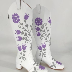 May include: A pair of white leather knee-high boots with a floral embroidery design. The flowers are purple and gray.