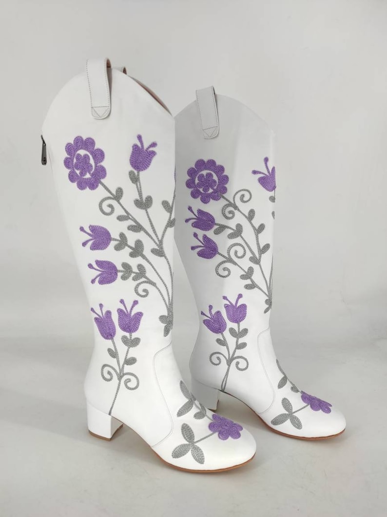 May include: A pair of white leather knee-high boots with a floral embroidery design. The flowers are purple and gray.