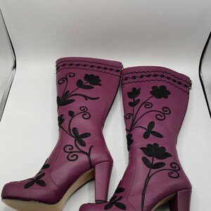 Puede incluir: Un par de botas hasta la rodilla de color ciruela con bordados florales negros que se extienden desde la punta hasta la parte superior de la ca&ntilde;a. Las botas tienen tac&oacute;n de bloque, punta redondeada y cierre de cremallera en la pantorrilla.