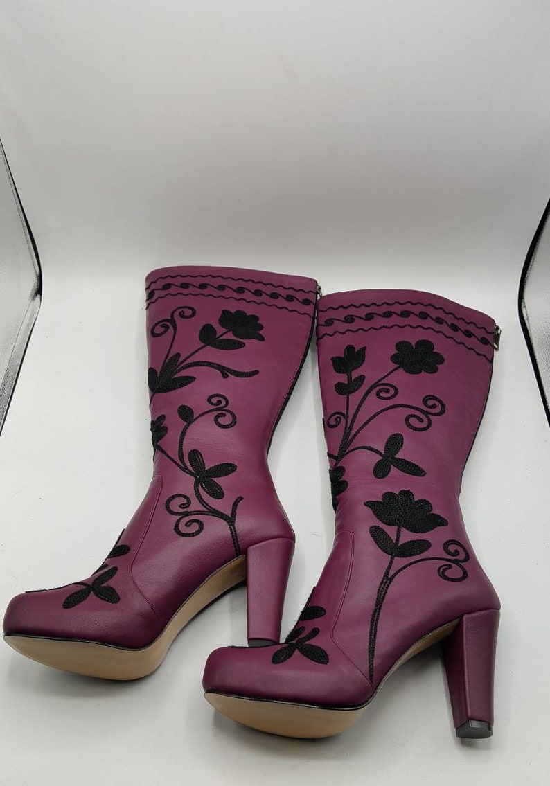 Puede incluir: Un par de botas hasta la rodilla de color ciruela con bordados florales negros que se extienden desde la punta hasta la parte superior de la ca&ntilde;a. Las botas tienen tac&oacute;n de bloque, punta redondeada y cierre de cremallera en la pantorrilla.