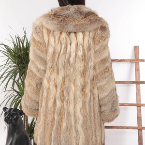 May include: A long, light brown fur coat with a wide collar. The coat has a vertical striped pattern and is worn by a person. The background includes a plant and a wooden ladder.