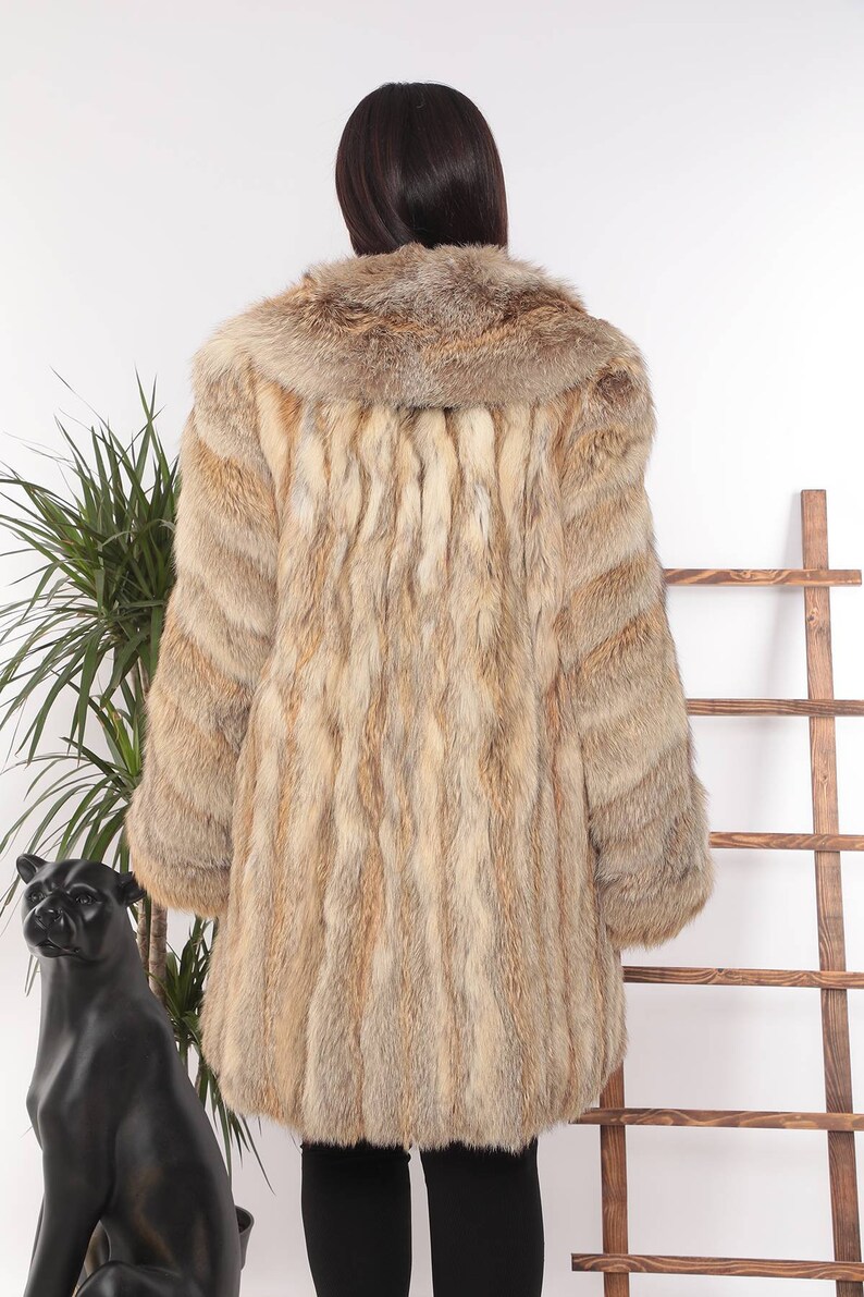 May include: A long, light brown fur coat with a wide collar. The coat has a vertical striped pattern and is worn by a person. The background includes a plant and a wooden ladder.