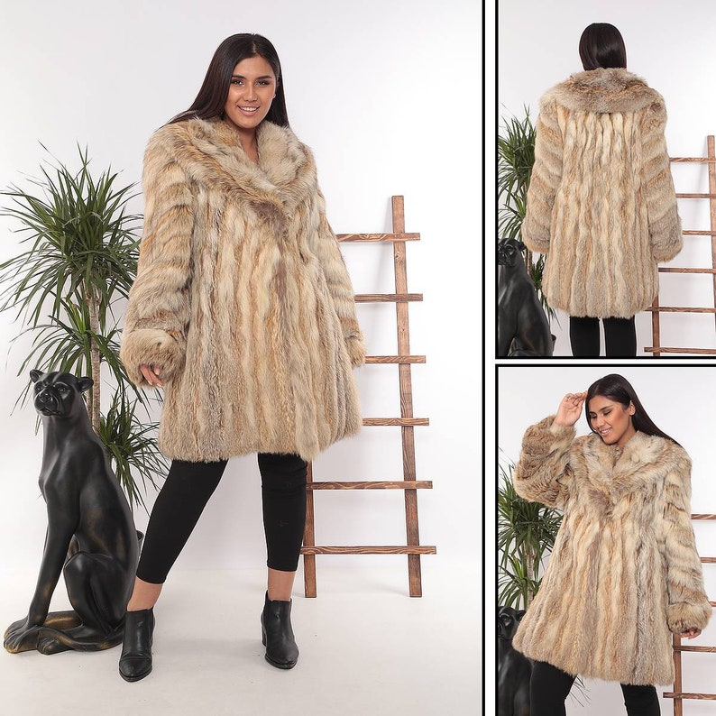 May include: A long, light brown fur coat with a wide collar and long sleeves. The coat has a vertical striped pattern and a slightly flared shape. The model is wearing black leggings and black boots.