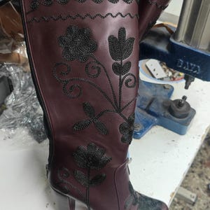 Puede incluir: Una bota alta de cuero color burdeos con patrones florales bordados en negro y costuras onduladas decorativas en la parte superior. La bota incluye una cremallera lateral y un tac&oacute;n de bloque con detallado trabajo de hilo negro.