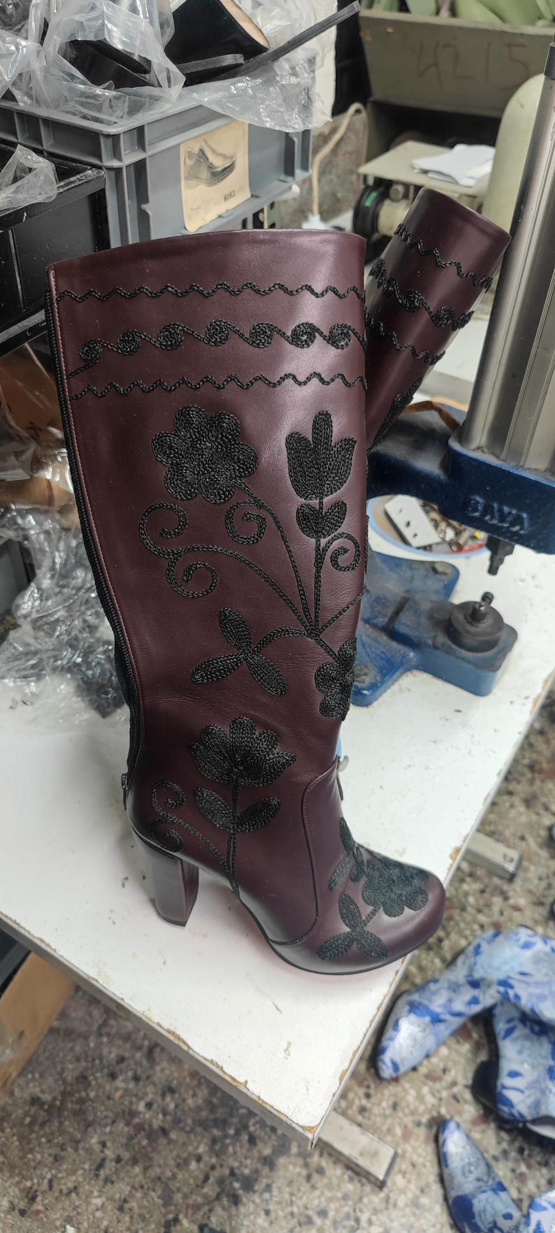Puede incluir: Una bota alta de cuero color burdeos con patrones florales bordados en negro y costuras onduladas decorativas en la parte superior. La bota incluye una cremallera lateral y un tac&oacute;n de bloque con detallado trabajo de hilo negro.