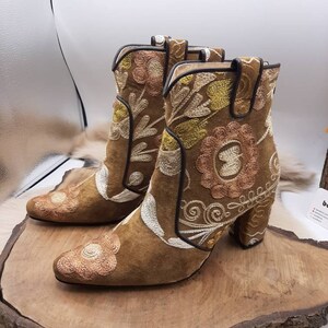 May include: Brown suede ankle boots with embroidered floral patterns in white, yellow, and pink. The boots have a block heel.