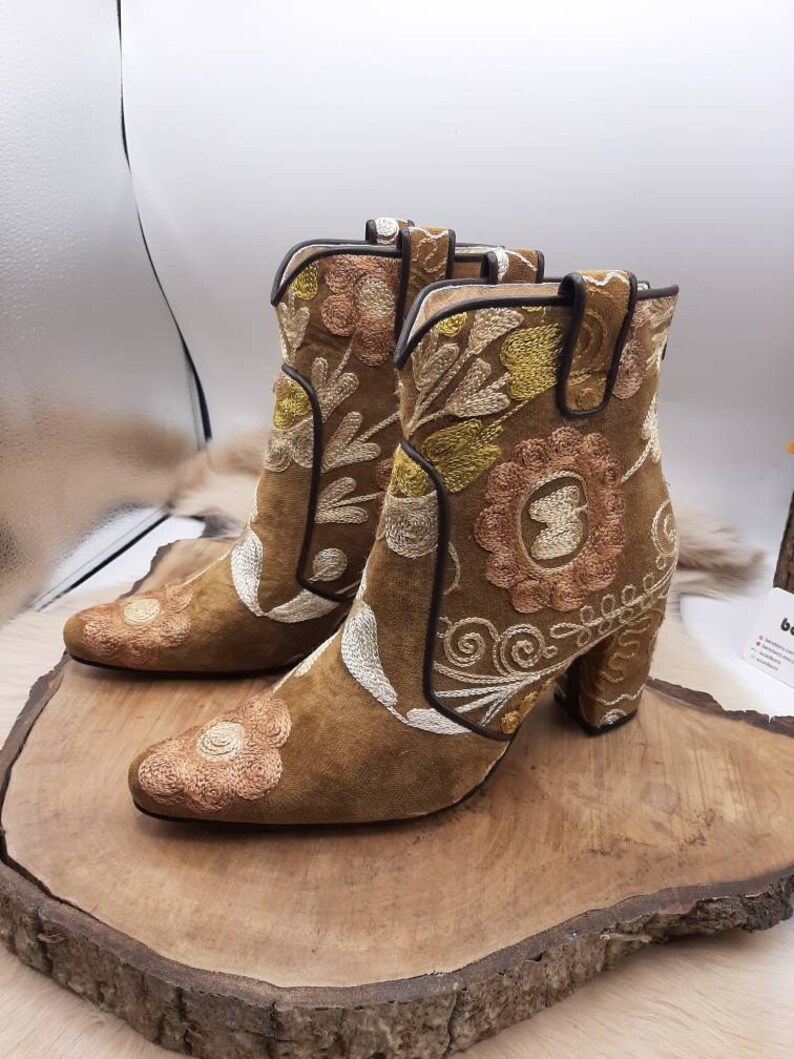 May include: Brown suede ankle boots with embroidered floral patterns in white, yellow, and pink. The boots have a block heel.