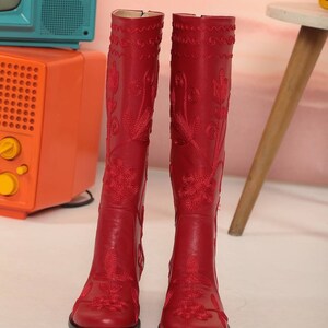 Puede incluir: Botas rojas hasta la rodilla con intrincado bordado floral. Las botas tienen punta y un tac&oacute;n peque&ntilde;o. El bordado es de un rojo m&aacute;s oscuro, creando un hermoso contraste. Las botas est&aacute;n hechas de un material liso similar al cuero.