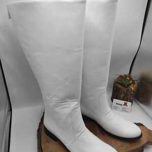 Puede incluir: Un par de botas blancas de cuero hasta la rodilla con tac&oacute;n bajo. Las botas est&aacute;n sobre una superficie de madera.