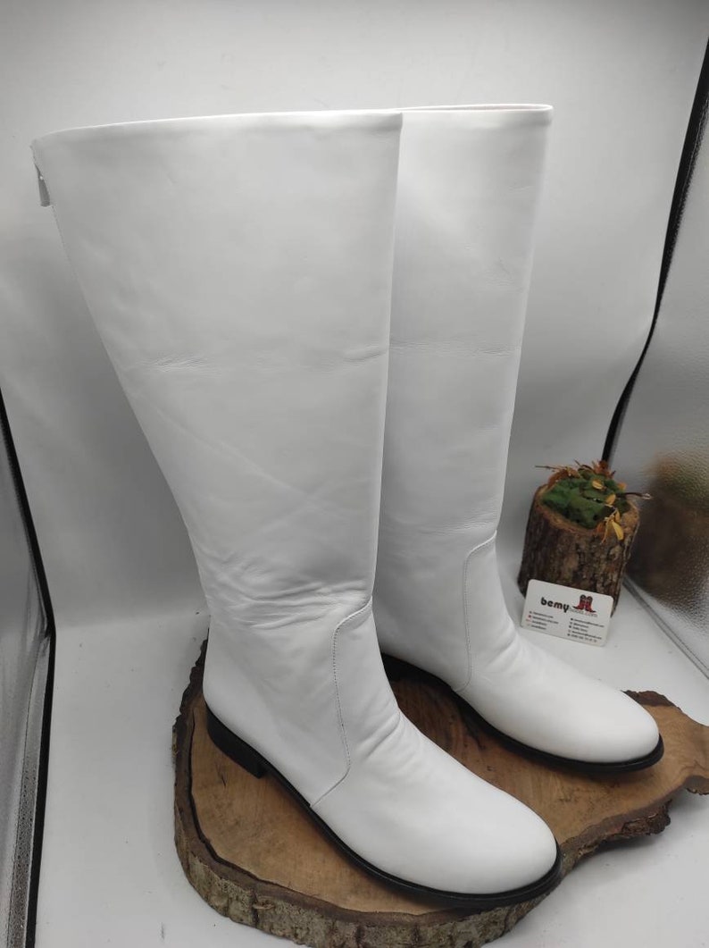 Puede incluir: Un par de botas blancas de cuero hasta la rodilla con tac&oacute;n bajo. Las botas est&aacute;n sobre una superficie de madera.