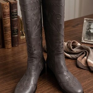 Puede incluir: Botas altas de cuero gris con intrincado bordado floral. Las botas tienen punta redonda y tac&oacute;n bajo. El dise&ntilde;o incluye costuras decorativas en el borde superior y en la parte delantera. Miden aproximadamente 40 cm de alto.