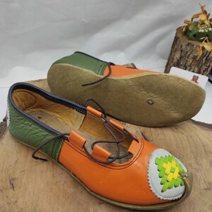 K&ouml;nnte beinhalten: Ein Paar orangefarbene und gr&uuml;ne Lederschuhe mit braunen Schn&uuml;rsenkeln. Die Schuhe haben ein dekoratives Design an der Spitze.